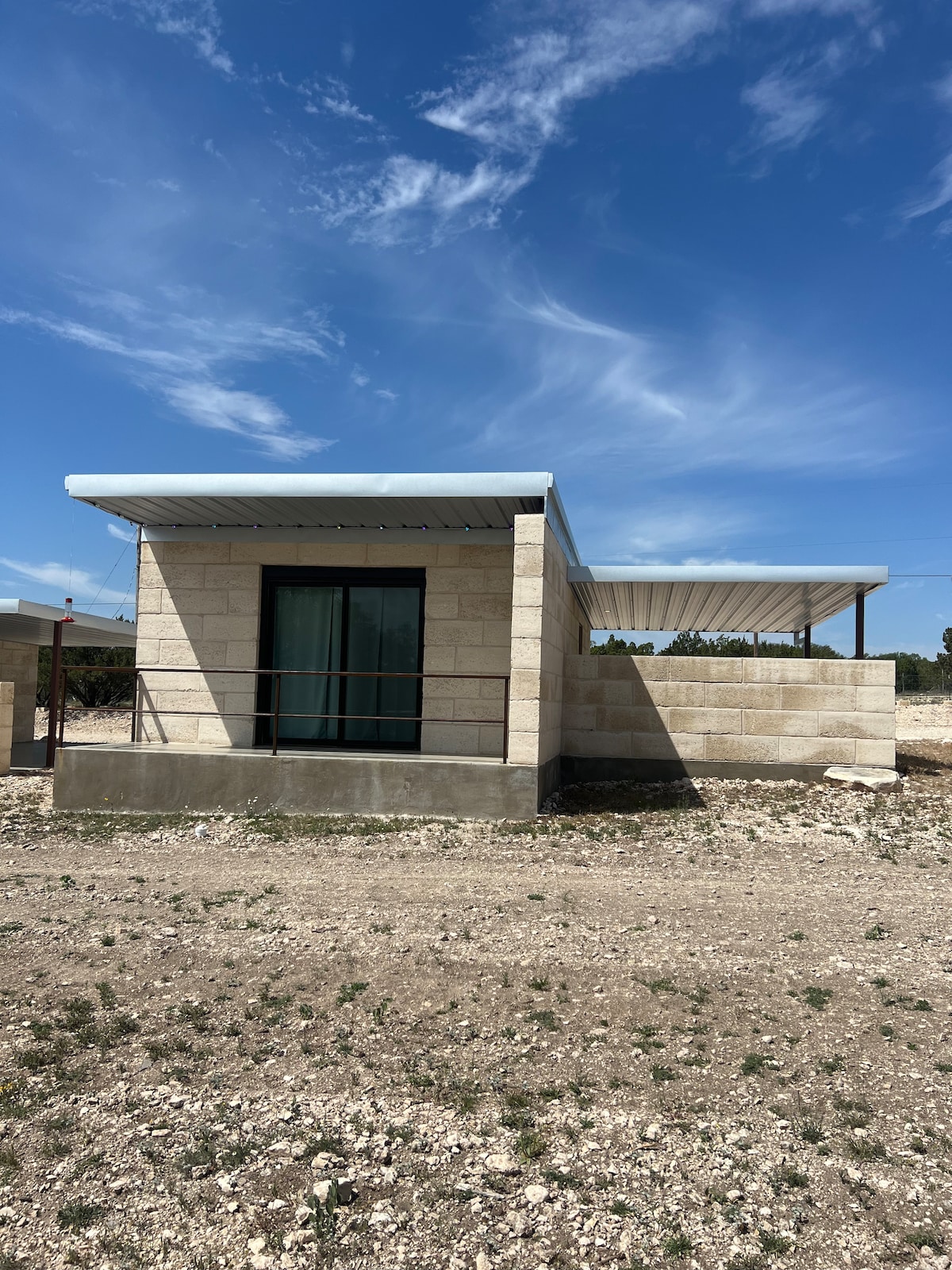 A limestone building is pictured against a clear blue sky, featuring a covered porch area. Large windows provide natural light, while a gravelly landscape surrounds the structure. The minimalist design reflects a modern approach, with a functional layout that harmonizes with the surrounding environment.