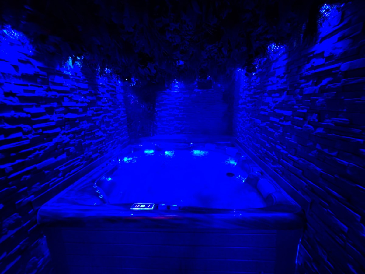 A spacious jacuzzi area is illuminated with soft blue lighting, creating an intimate ambiance. Stone walls surround the jacuzzi, while subtle LED lights enhance the soothing environment. Control panels for temperature and settings are visible on the edge of the unit.