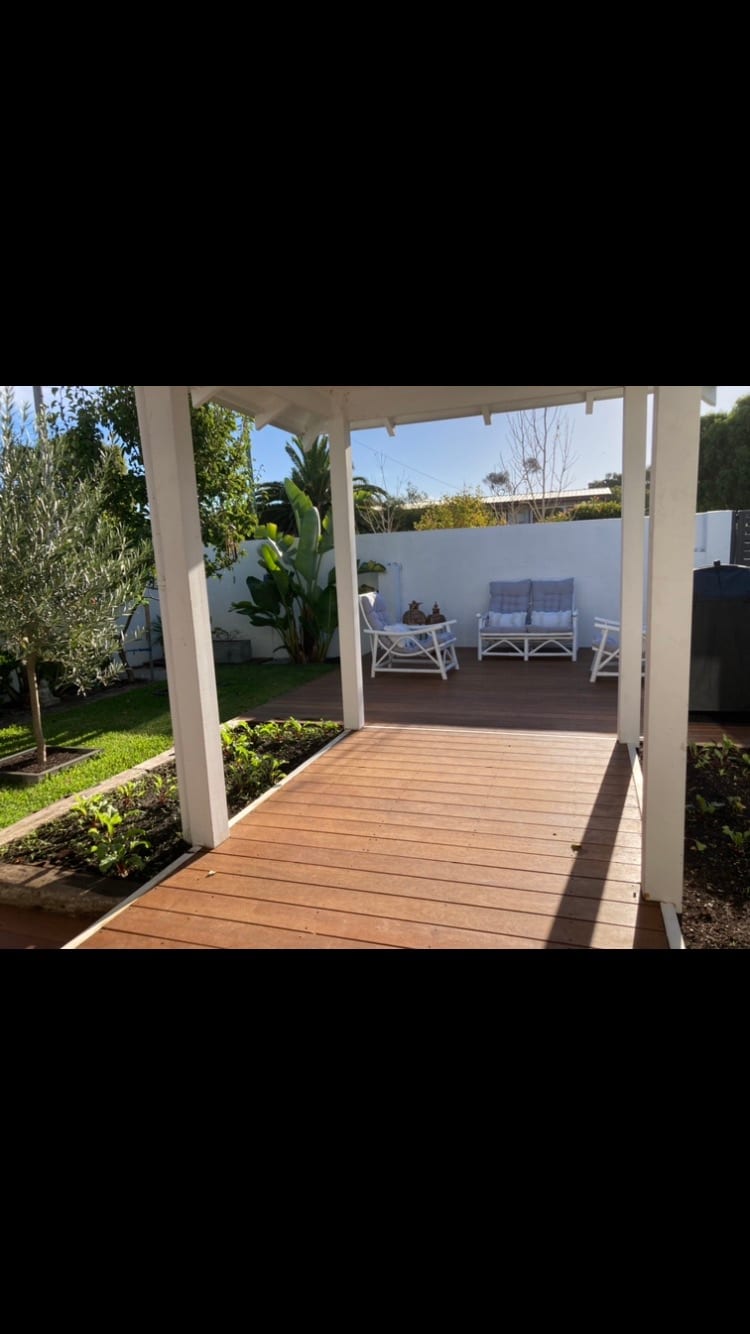 A wooden walkway leads into a peaceful courtyard, surrounded by greenery. In the background, a pair of white chairs and a small table invite relaxation. The area is bathed in natural light, enhancing the tranquil atmosphere of the outdoor space.