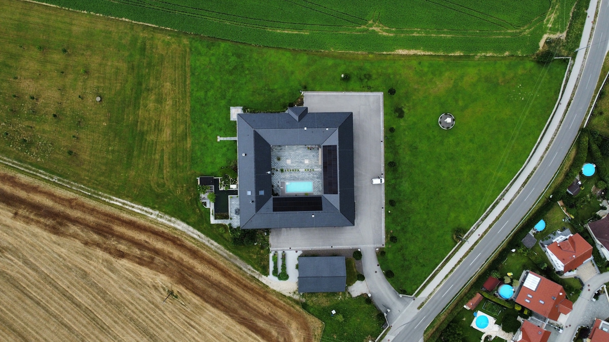 An aerial view of the property reveals a large building with a modern design, surrounded by green lawns. A pool is visible at the center, while the surrounding area includes neatly maintained fields and a winding road. Nearby residences are seen in the distance.