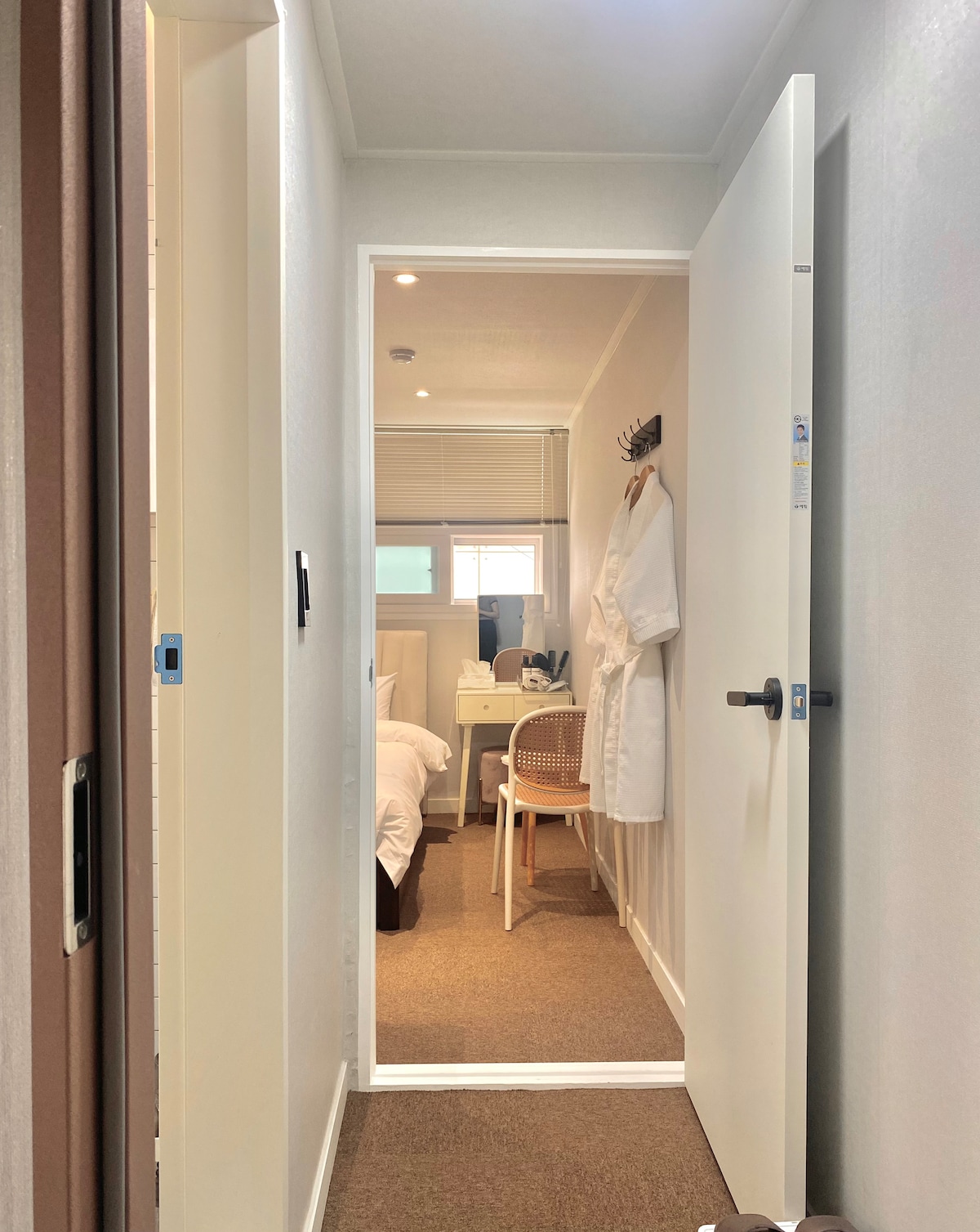 A hallway leads to a room featuring warm lighting. A chair and small table are visible, along with a bathrobe hanging on the wall. The slightly ajar door provides a glimpse of the sleeping area, which includes a bed and a window.