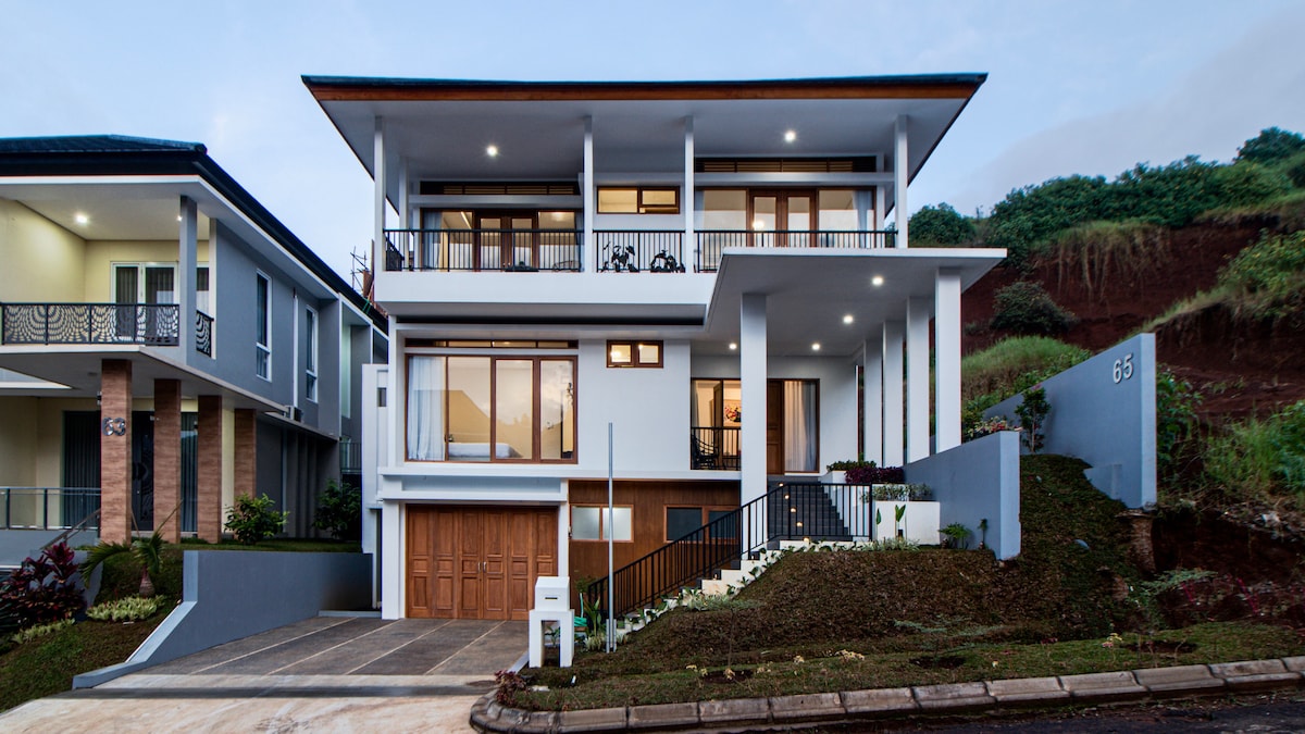 A modern villa is presented with a clean architectural design, showcasing multiple large windows that invite natural light. The exterior features a combination of white walls and a wooden garage door, complemented by neatly landscaped greenery and a sloped pathway leading to the entrance.