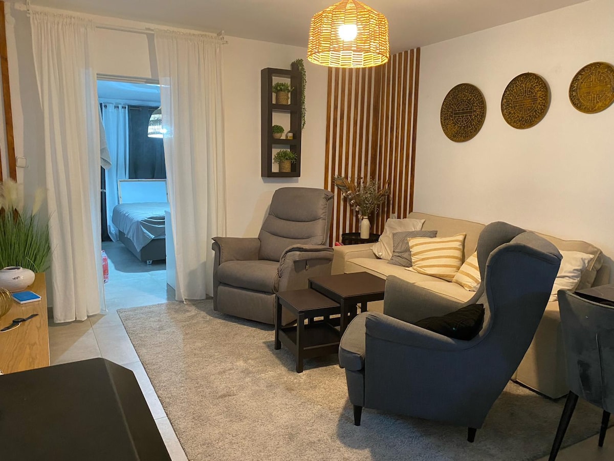 A living area is featured with comfortable furniture, including two armchairs and a sofa. A coffee table sits on a soft area rug. Decorative wall art and a light fixture enhance the space. Light from an adjacent room adds brightness to the atmosphere.