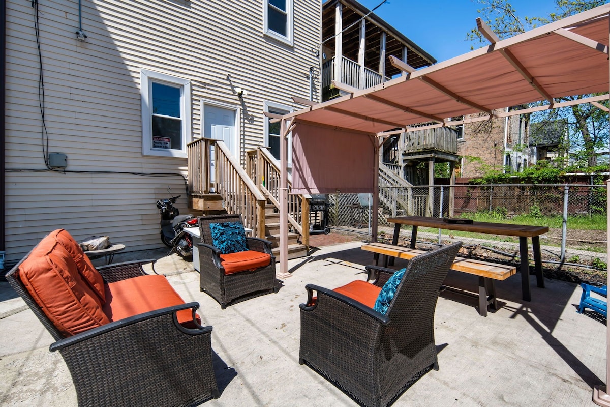 A spacious outdoor area features a shaded gazebo with seating and a table. Two comfortable chairs with orange cushions are positioned next to the table, surrounded by a concrete patio and a fenced area. Stairs lead to the entrance of the building.