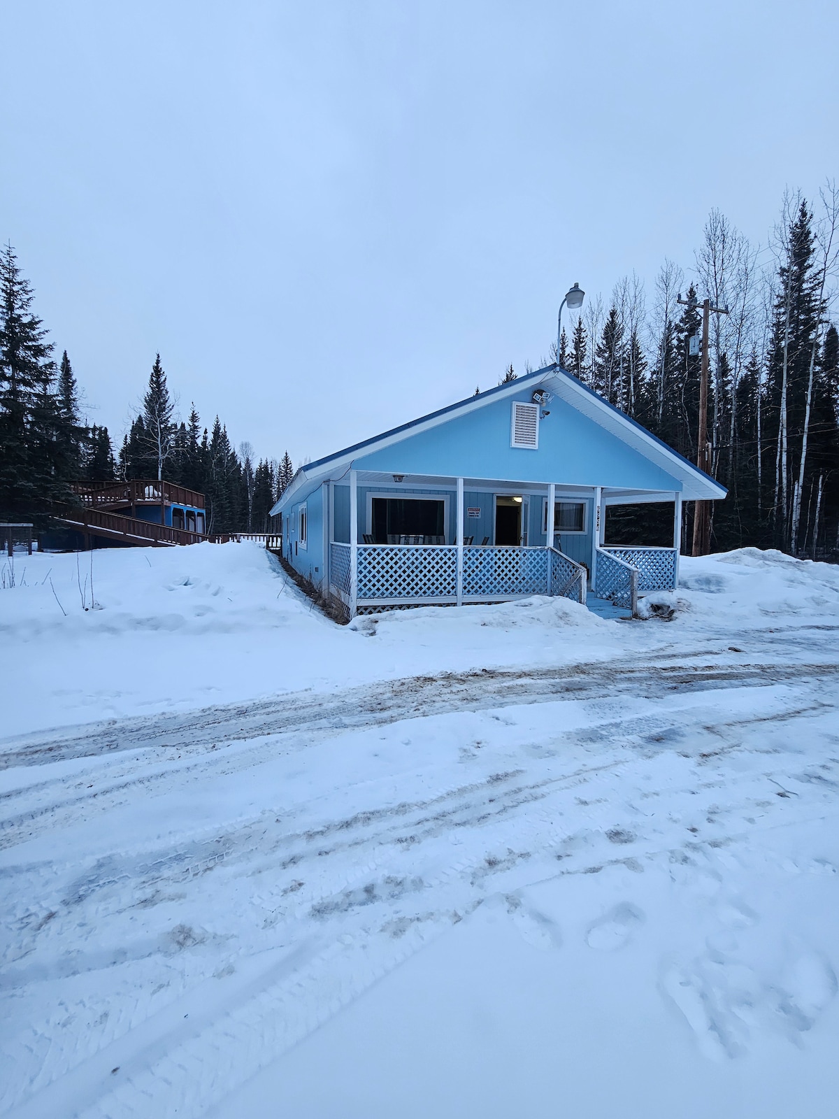 A light blue house is framed by snow-covered ground and tall trees, creating a serene winter landscape. The front porch is accessible via a gentle ramp, and the driveway shows evidence of winter travel.