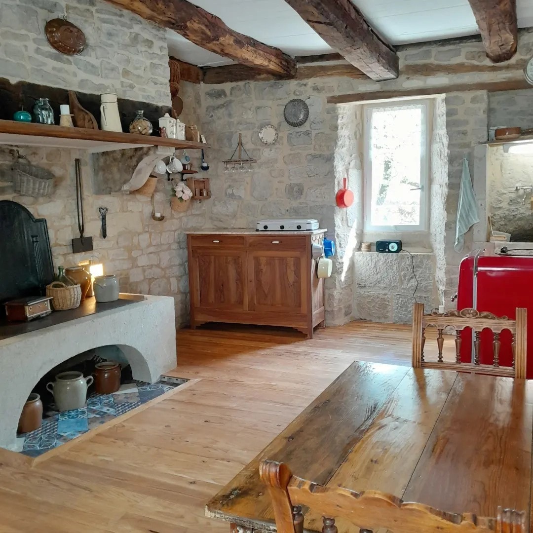 A rustic kitchen area features exposed stone walls and wooden beams. A wooden table is placed centrally, complemented by wooden chairs. A vintage red refrigerator and a classic stove are positioned against the wall, while decorative items adorn the shelves and fireplace.