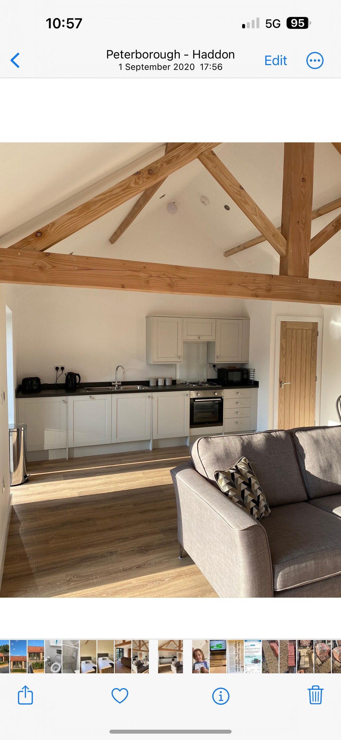 An open plan living area features vaulted ceilings supported by wooden beams. A light-colored kitchen with a modern finish is visible, accompanied by black appliances. Sunlight streams through large windows, illuminating the cozy seating area that includes a sofa adorned with decorative cushions.