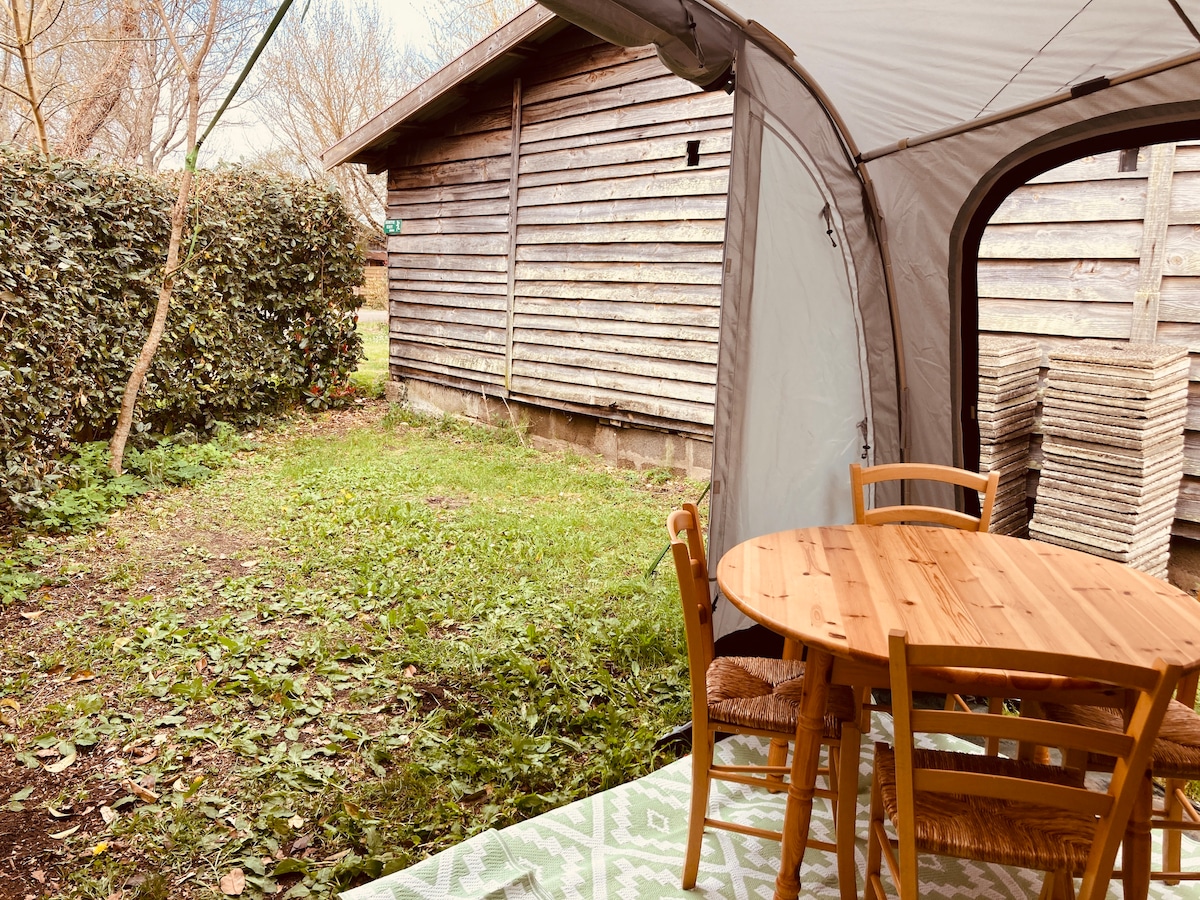 An outdoor seating area features a round wooden table accompanied by two chairs and one stool. The area is surrounded by green grass and shrubs, with a wooden structure in the background. Natural light filters through the space, creating a serene ambiance.