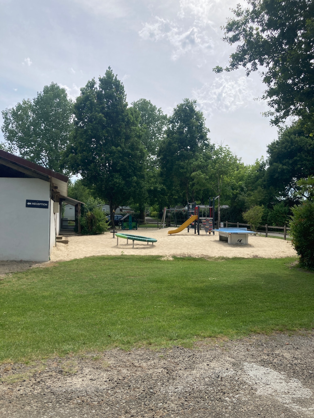 A sandy playground area is surrounded by grassy space, featuring a slide and climbing structure for children. The scene includes several trees providing shade, and facilities are accessible nearby, adding to the inviting atmosphere of the camping environment.