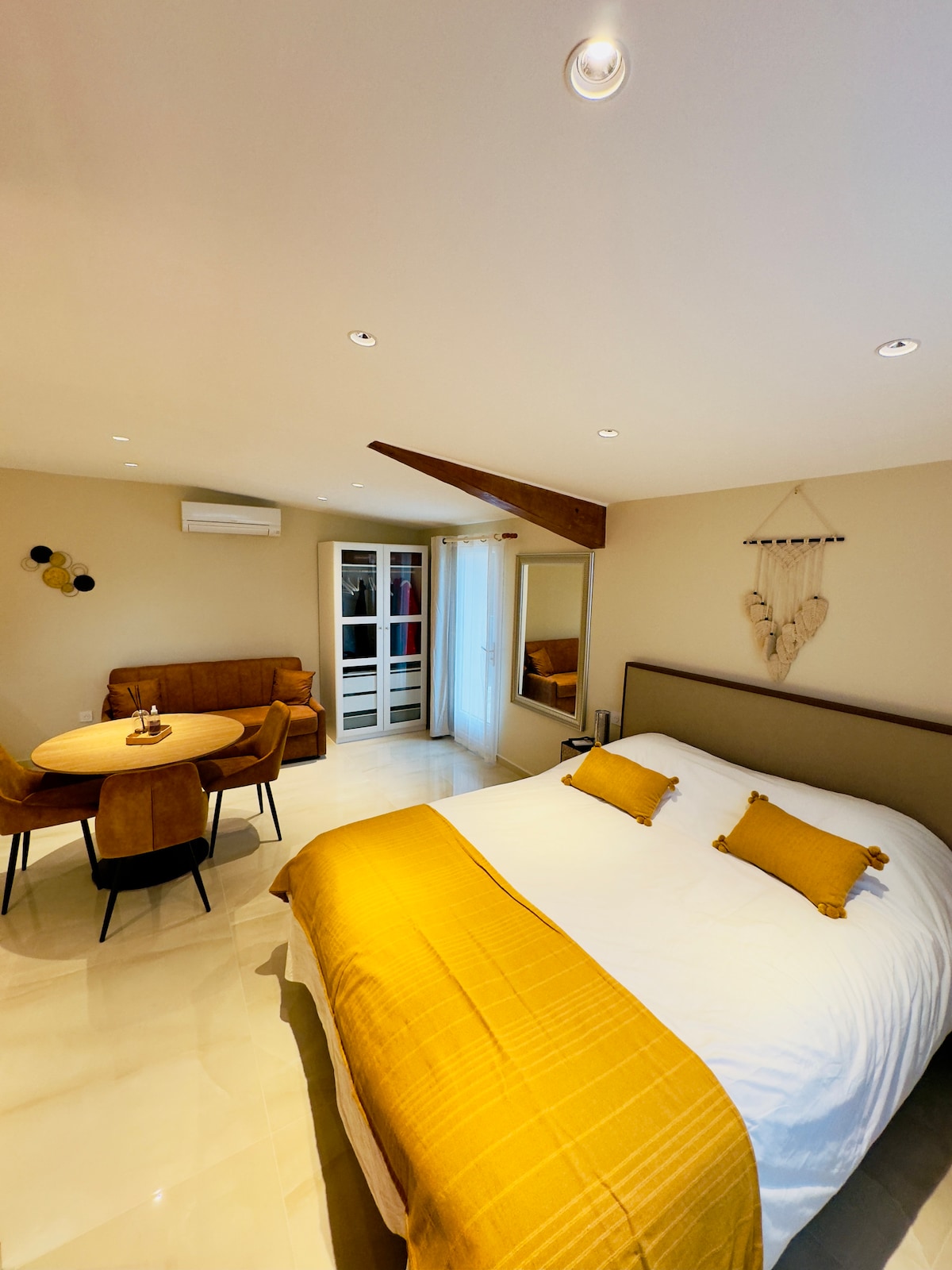 A spacious sleeping area features a large bed adorned with two yellow accent pillows. A cozy sitting area includes a brown sofa and a round dining table with wooden chairs. Natural light enters through glass doors, enhancing the room's open feel.