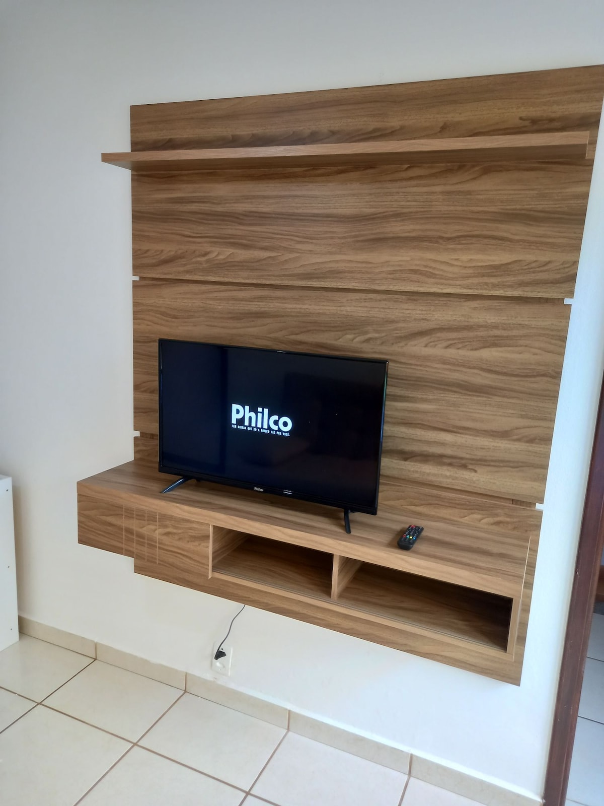 A modern wooden wall unit is featured, supporting a flat-screen television. Below the TV, an open shelf is available for storage. The flooring is tiled, and neutral walls complement the natural wood tones, contributing to a simple and functional design.