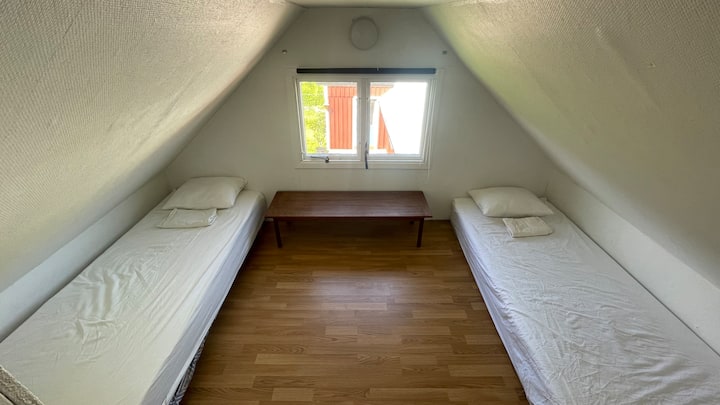 Dhomë gjumi e rehatshme në katin e sipërm me tavane të pjerrëta, dy krevate tekë, një hyrje me perde për privatësi dhe pamje të kopshtit të pasmë.
