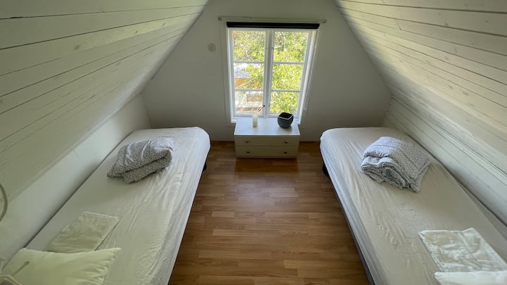 Dhomë gjumi e ndritshme në katin e sipërm me tavane të pjerrëta, dy krevate tekë, një hyrje me perde për privatësi dhe pamje panoramike nga oborri anësor.