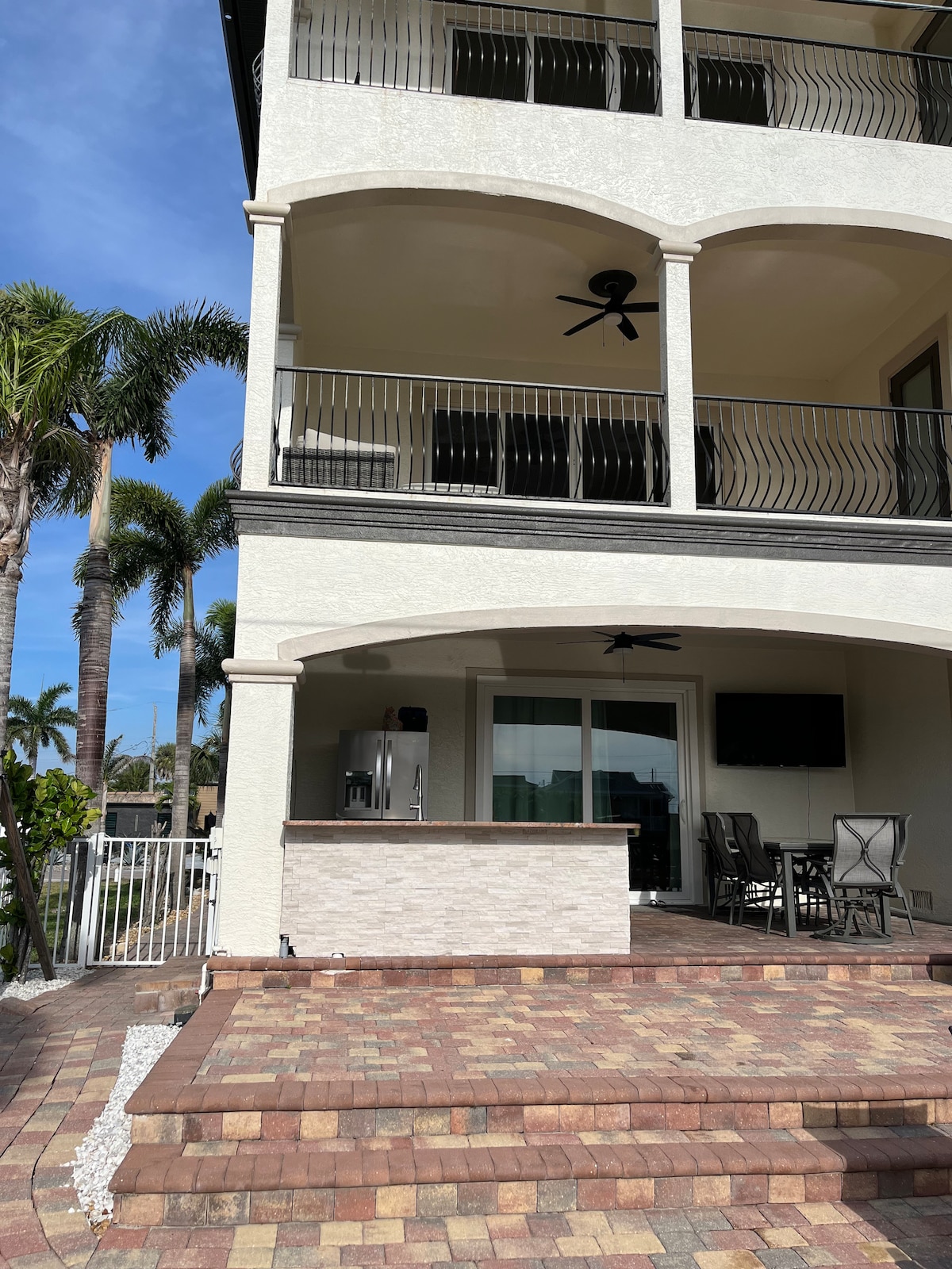The exterior of a multi-story waterfront home is showcased, featuring a spacious patio with multiple seating options. A barbecue grill is visible, along with a dining area. Decorative brick paving leads up to the entrance, while palm trees provide a tropical touch to the environment.