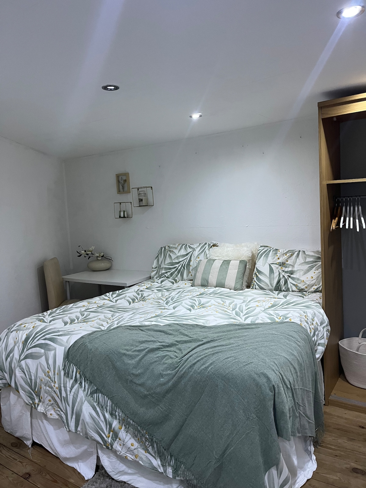 A comfortable sleeping area features a spacious bed adorned with a botanical-patterned duvet and a coordinating green throw blanket. Soft lighting is provided by recessed ceiling fixtures. A small side table and an open closet with hangers are visible, enhancing the room's functionality.
