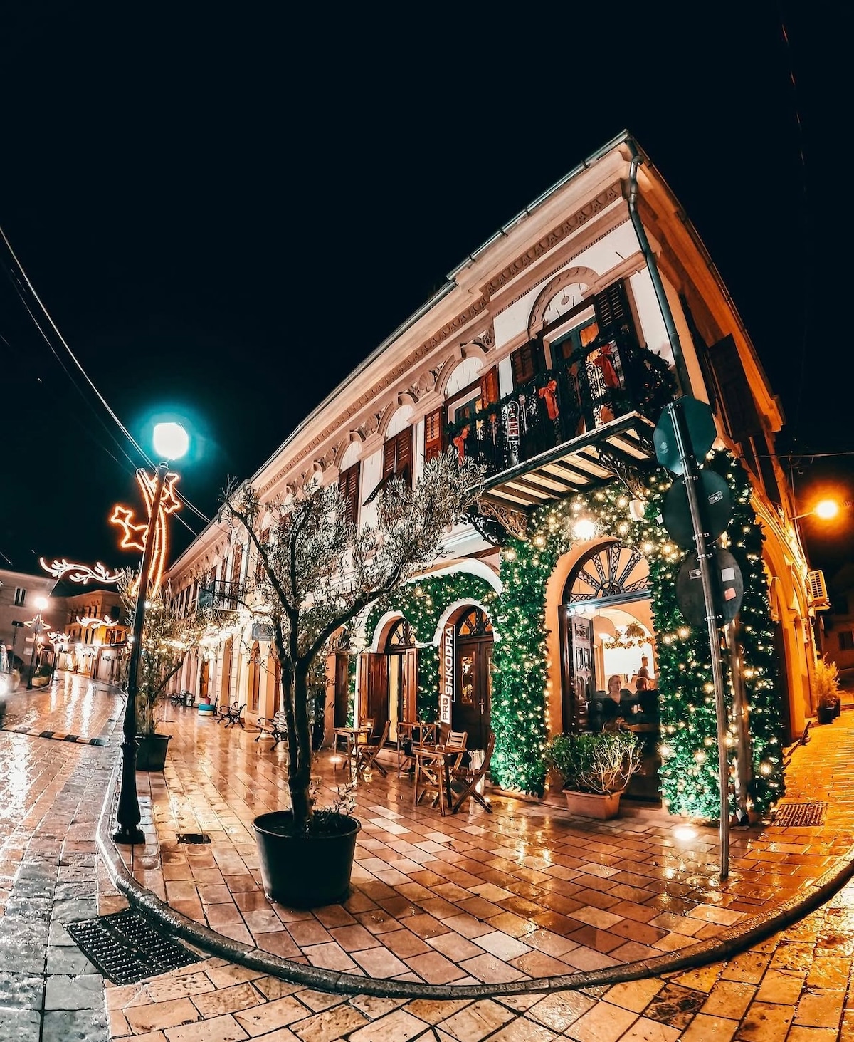 A charming corner building is illuminated with festive lights, surrounded by greenery and outdoor seating. The wet pavement reflects the warm glow from the street lamps, and decorative elements enhance the inviting ambiance of the area at night.