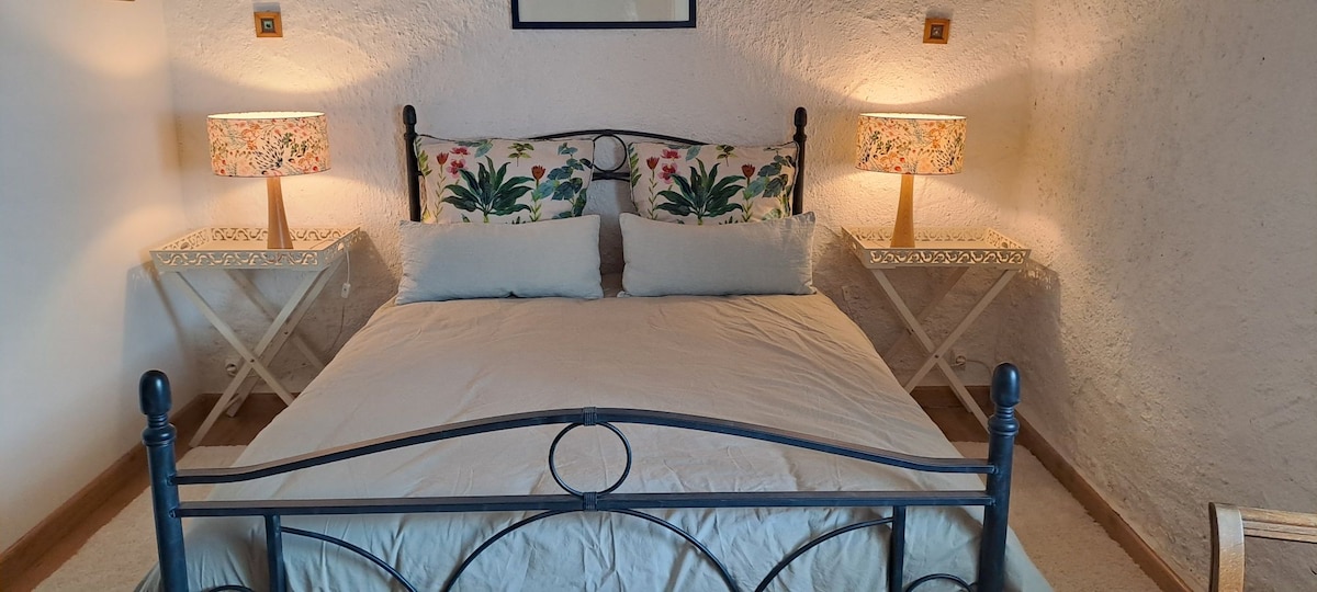 A cozy sleeping area features a dark metal bed frame with soft, light bedding. Two bedside tables with floral lamps provide gentle lighting, while artwork hangs above the headboard, adding a touch of character to the room.