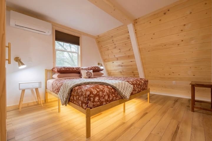King sized bed with nightstands, sconce lighting, mini split AC and heating unit, full length mirror, closet, window and pull down blinds, and on suite bathroom with sliding barn door