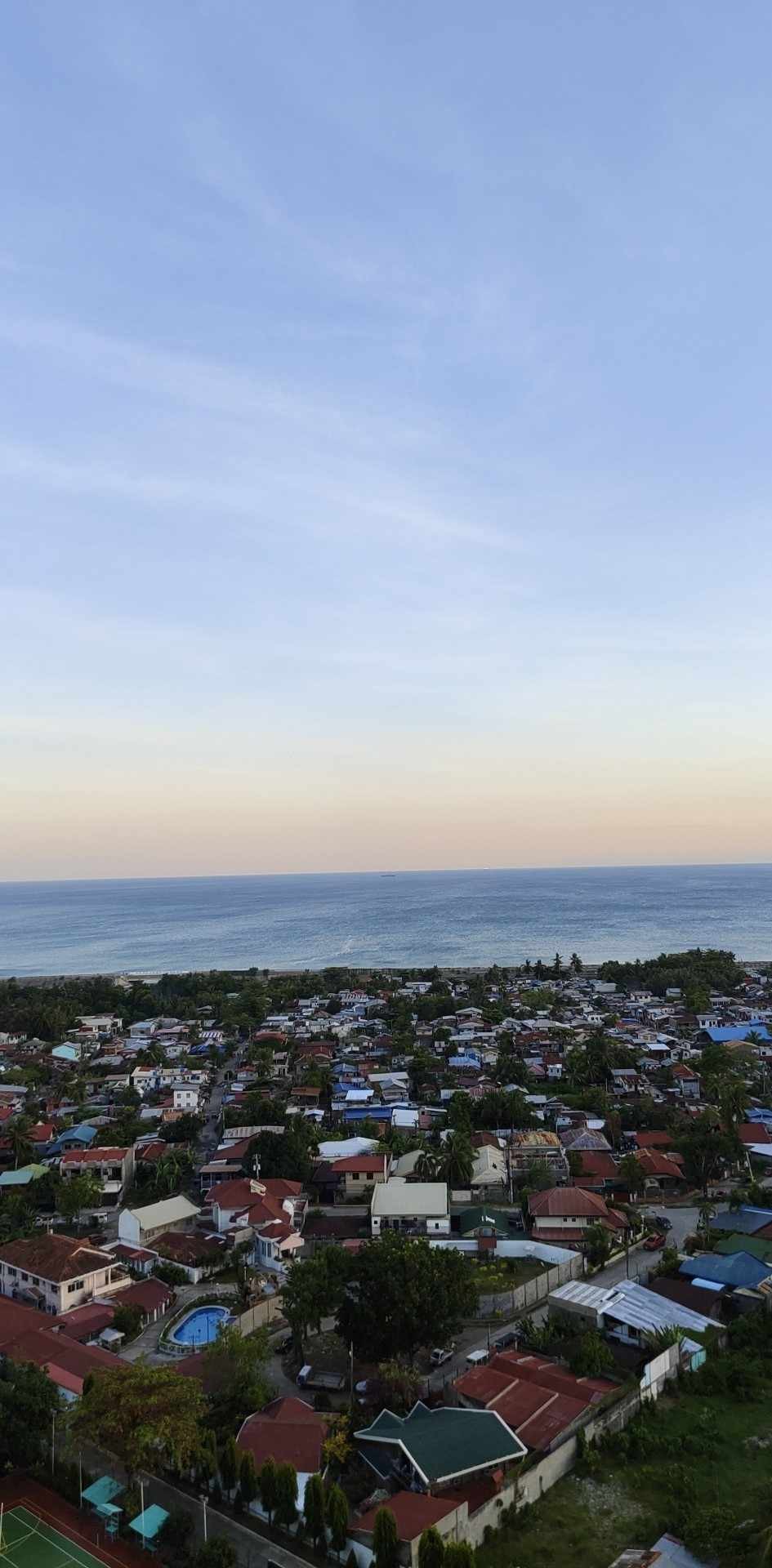 A panoramic view of the coastal landscape reveals a patchwork of houses with varying rooftops, transitioning from inland greenery to the open sea. The horizon features soft hues of blue and orange, suggesting a serene sunset over the water.