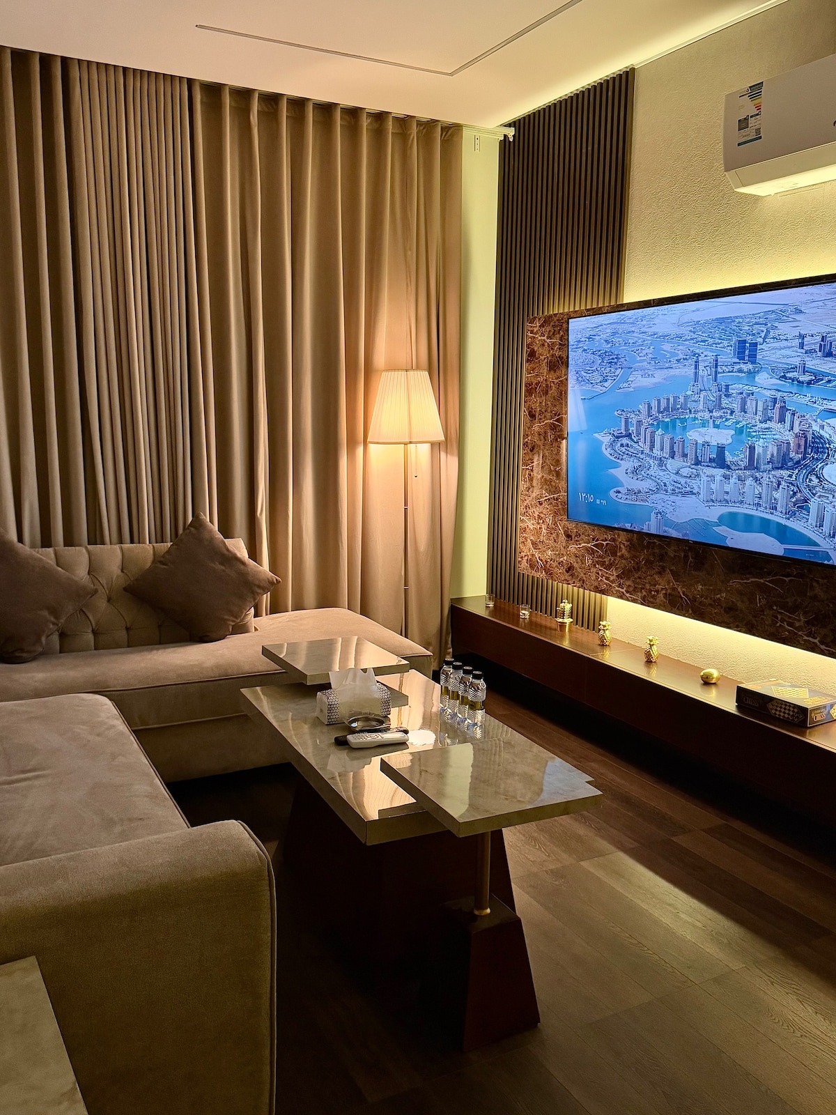 A modern living area includes a plush beige sofa and a decorative coffee table. A smart TV is mounted on the wall, showcasing a cityscape. Soft lighting is provided by a floor lamp, and elegant curtains frame the window, adding a touch of warmth to the space.