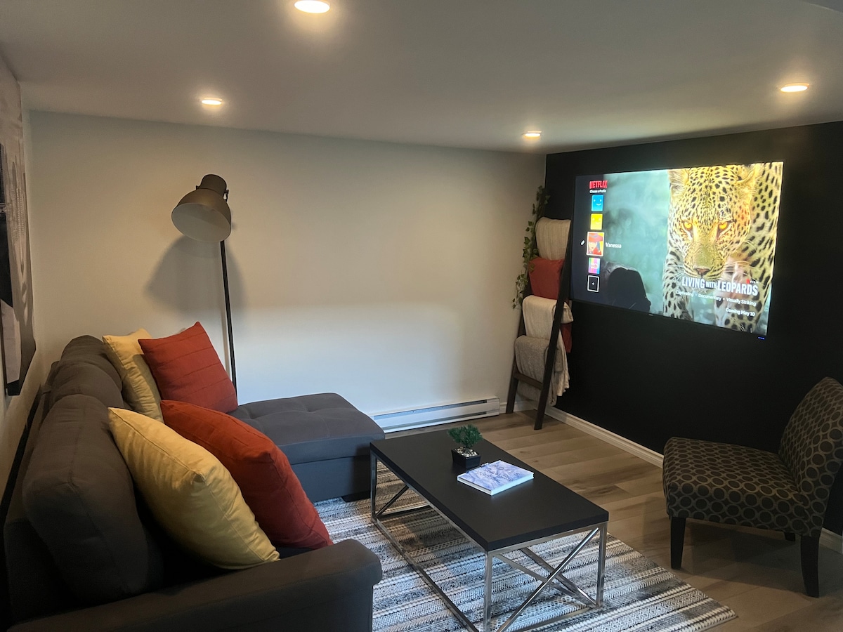 A cozy living area is featured with a grey sectional sofa adorned with colorful cushions. A coffee table is positioned in front, complementing a flat-screen TV mounted on the wall. Soft lighting enhances the modern decor and creates a comfortable space for relaxation.