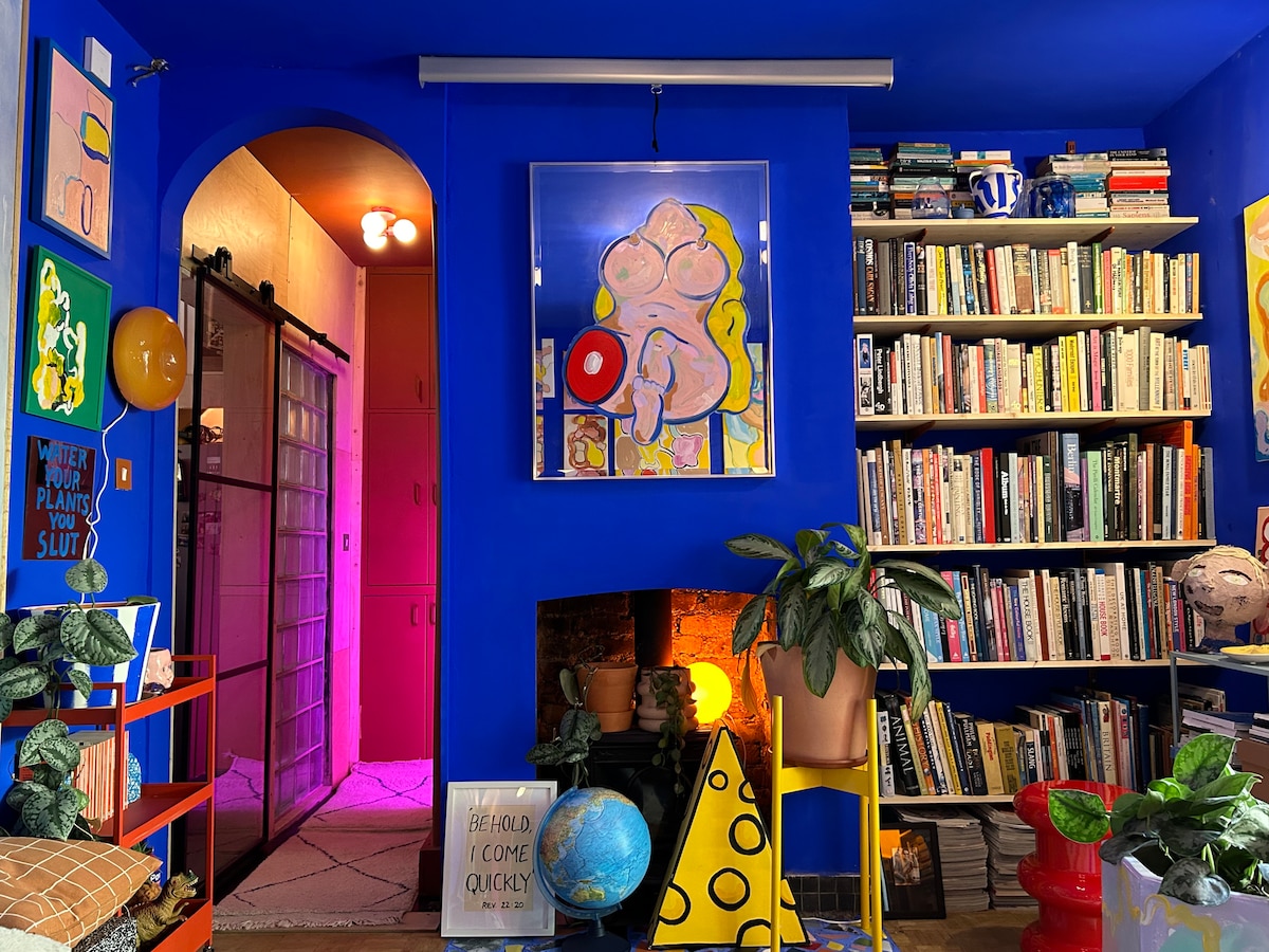 A vibrant living space features bold blue walls adorned with colorful art pieces. A bookshelf filled with books occupies one side, while a cozy fireplace is partially visible beneath artwork. A doorway leads to a warmly lit area, adding depth to the lively atmosphere.