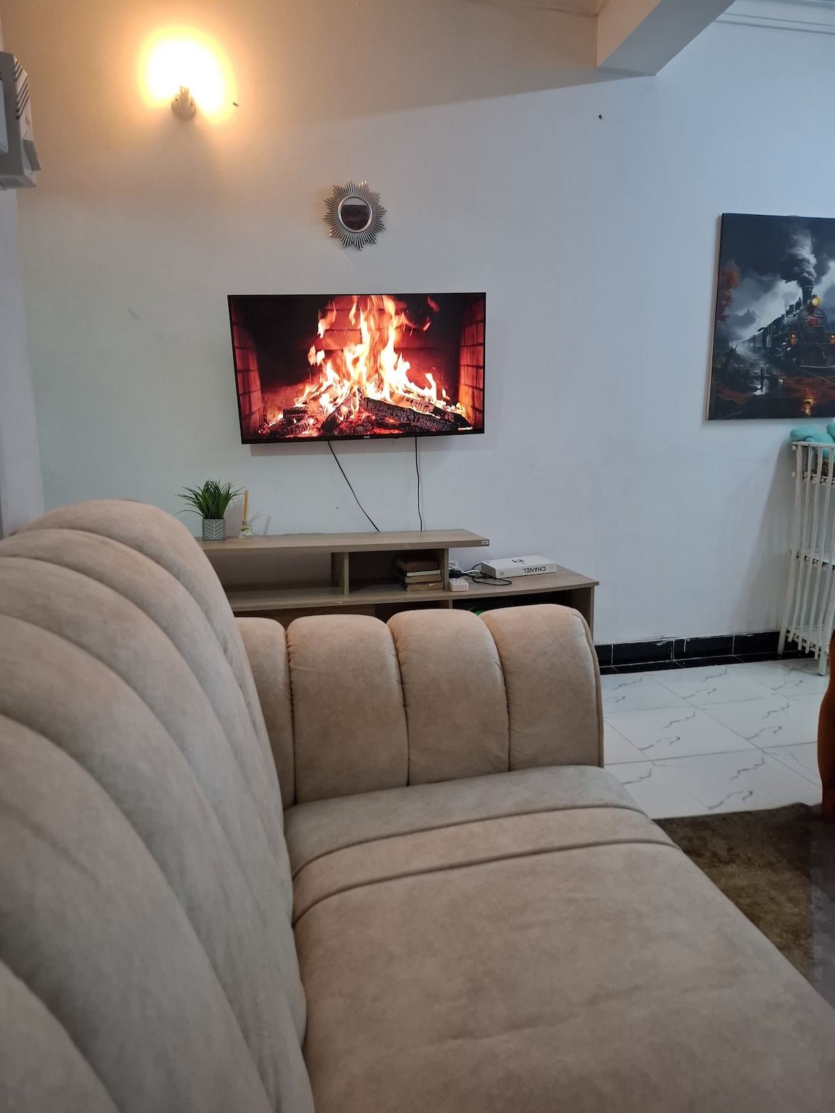 A comfortable beige sofa is positioned facing a wall-mounted television displaying a flickering fireplace. A small decorative plant sits on the media console below the TV. The room features bright lighting and a subtle artwork hanging on the adjacent wall.