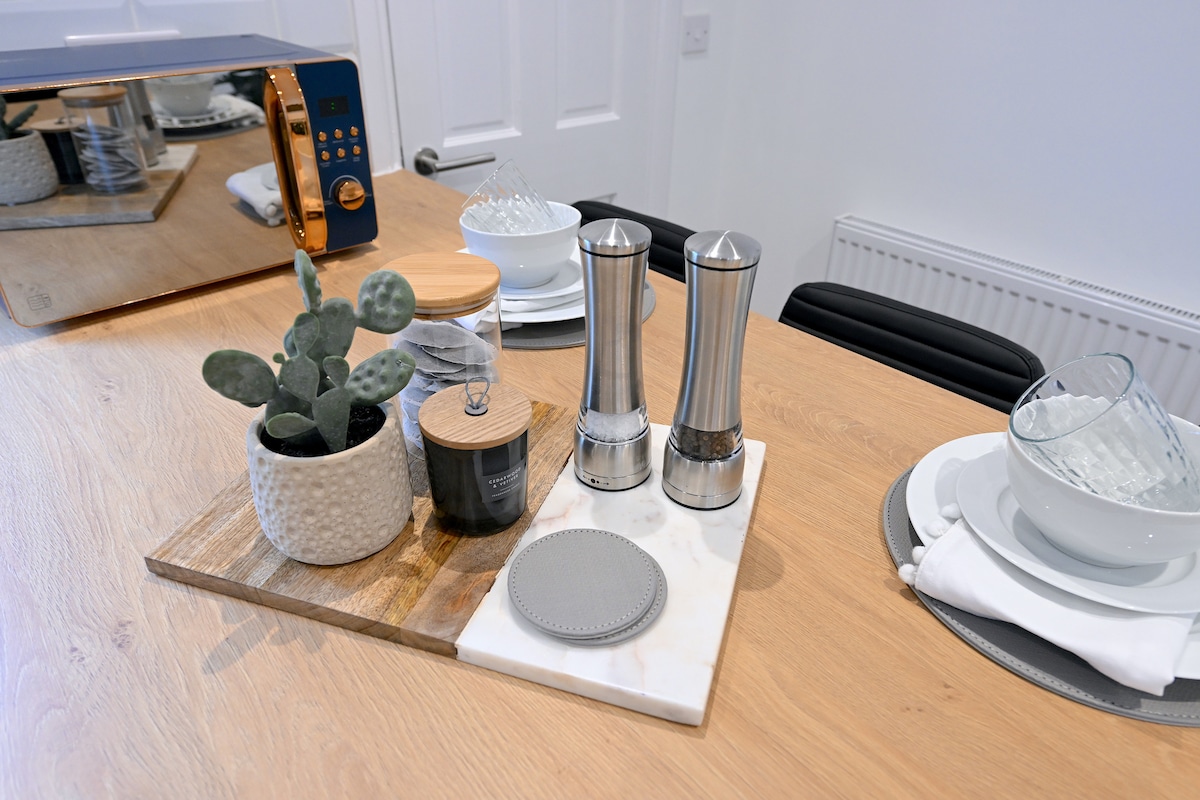 A neatly arranged kitchen countertop displays a selection of dining essentials, including stainless steel salt and pepper mills, a potted succulent, and a candle. Plates and bowls are set, complemented by a stylish placemat, contributing to a modern and organized atmosphere.