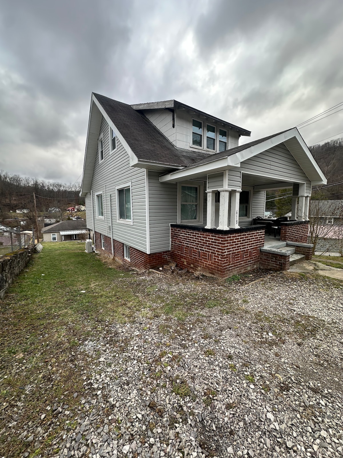The image captures a two-story house with a grey exterior, displaying a mix of siding and brick features. A covered porch is visible, with steps leading up to the entrance. The yard is grassy, bordered by gravel, with panoramic views of the surrounding area.