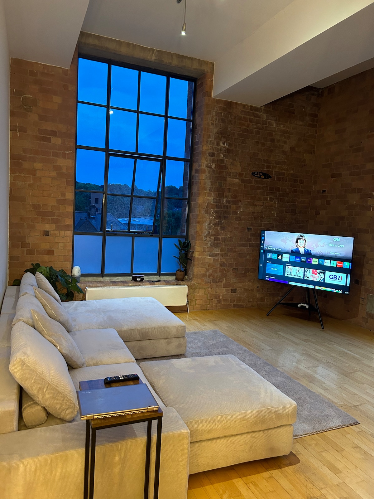 A spacious living area features a large sectional sofa in light fabric, positioned near a significant window with multiple panes. A sleek television stands on a stand opposite the seating area. Warm wooden flooring complements the exposed brick wall, enhancing the cozy atmosphere.
