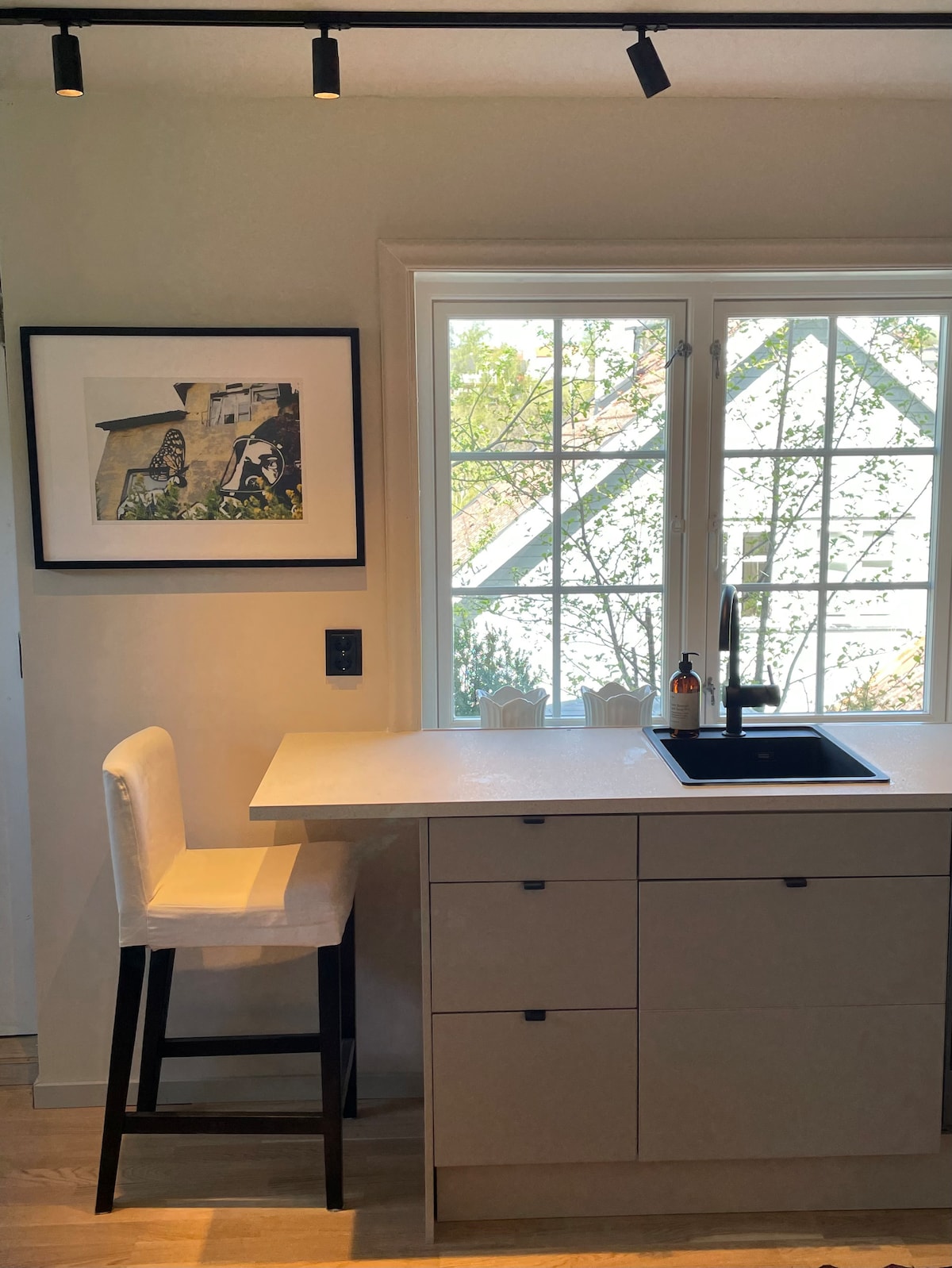 A modern kitchen is depicted, featuring a minimalist design with a light-colored countertop and cabinetry. A high stool sits next to the countertop, while a window allows natural light to illuminate the space, offering a view of greenery outside. A framed artwork hangs on the wall.