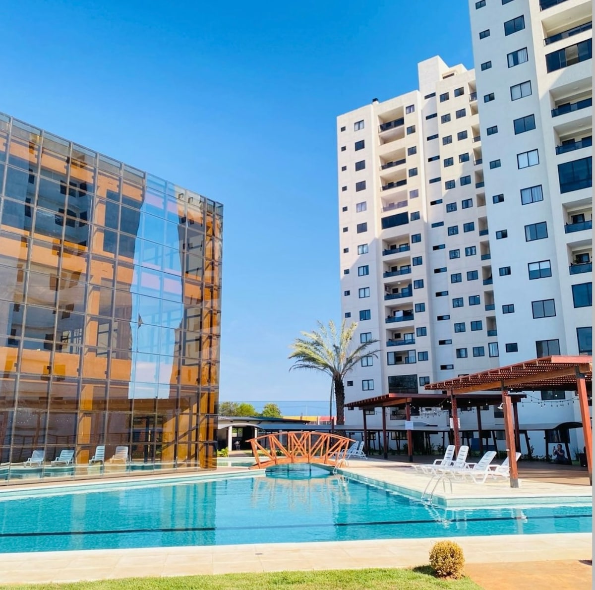 A modern swimming pool is surrounded by well-maintained greenery and lounge chairs. The pool reflects the clear blue sky, while contemporary apartment buildings flank one side, featuring a mix of glass and solid facades. Sunshade structures are visible near the pool area.