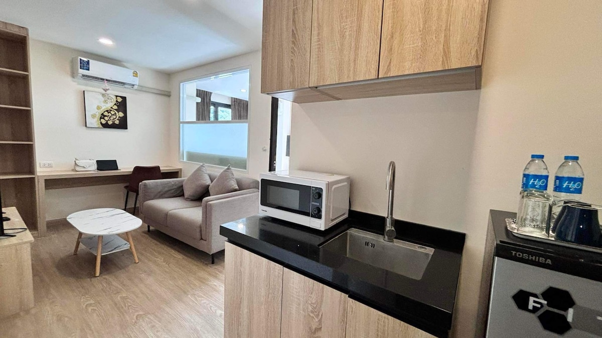 A compact kitchen area features modern wood cabinetry and a sleek black countertop. A microwave and sink are visible, accompanied by two bottles of water. A comfortable seating arrangement is indicated by a light-colored sofa facing a small coffee table, contributing to a functional living space.