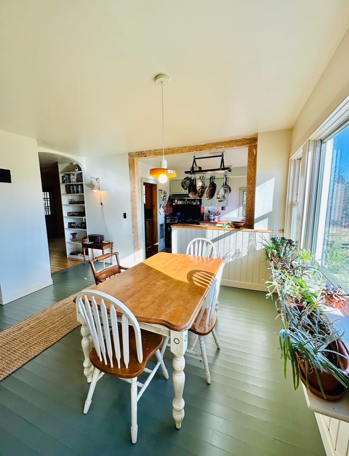 A bright dining area features a wooden table surrounded by four white chairs, with sunlight streaming through large windows. Hanging cookware is seen in the background, with plants lining the windowsill, adding a touch of greenery to the space.