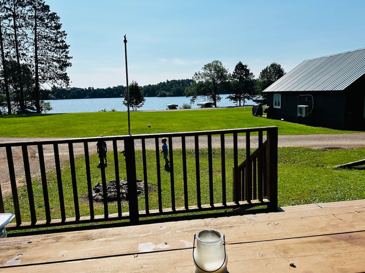 The outdoor deck overlooks a serene lake, with expansive green grass leading to the water's edge. A candle is placed on the wooden table in the foreground. A cabin can be seen to the right, framed by trees under a clear blue sky.