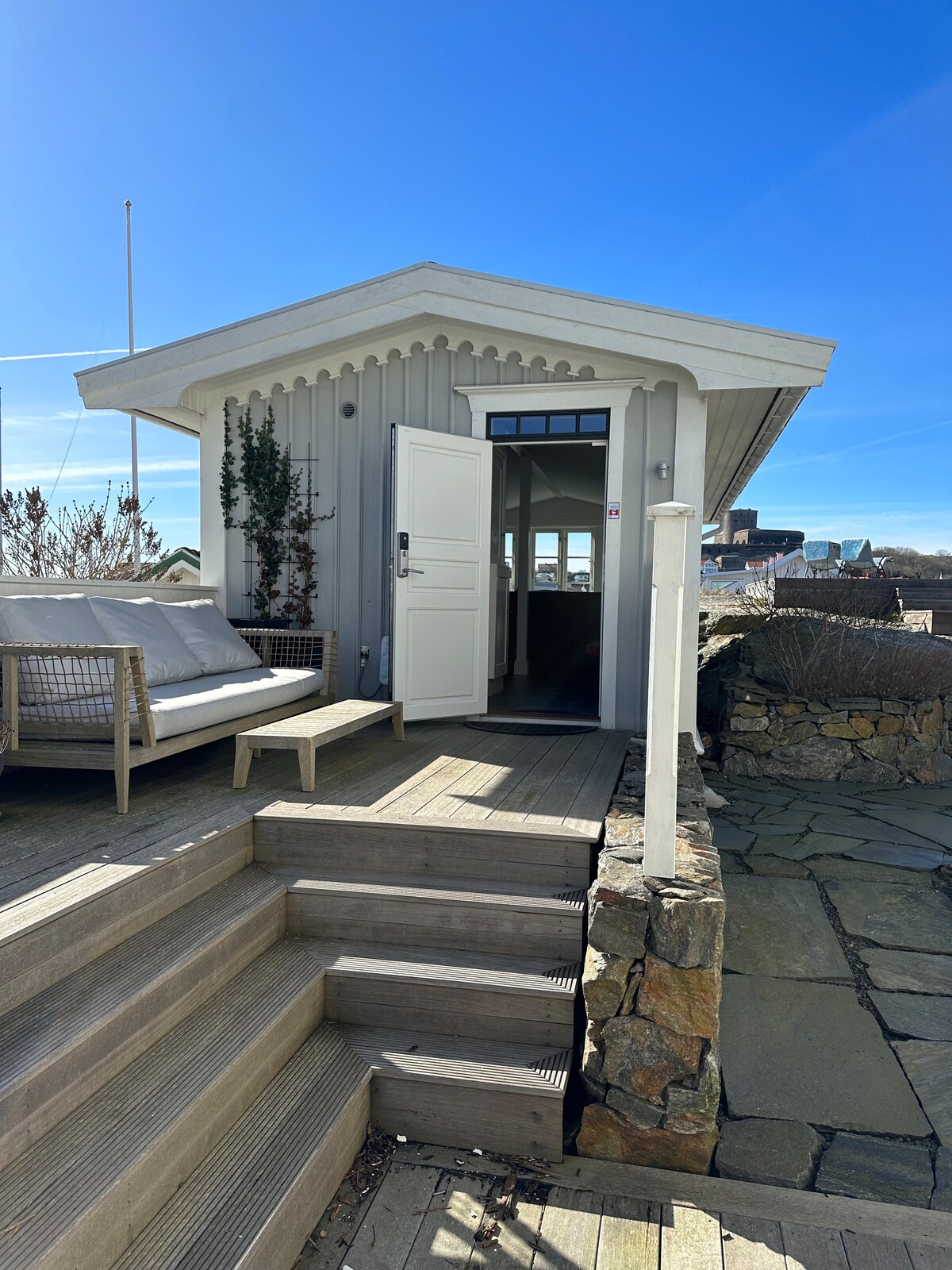 A charming entrance to a light-colored guesthouse is framed by a wooden deck. The door stands open, revealing a bright interior. Natural stone steps lead up to the entrance, surrounded by greenery and coastal scenery under a clear blue sky.