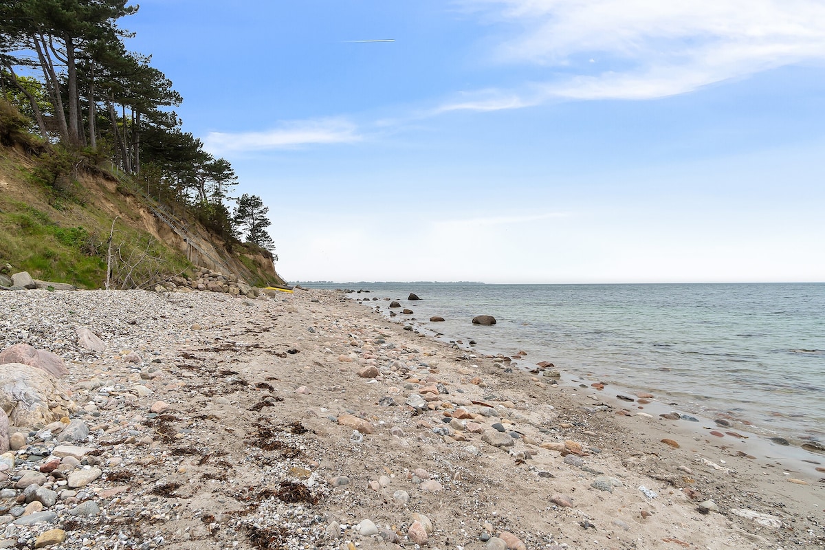 A serene beach scene is presented, showcasing a coastline with smooth pebbles and sand. Gentle waves lap at the shore, while a backdrop of trees rises along the cliff. The sky appears clear, enhancing the tranquil setting near the water's edge.