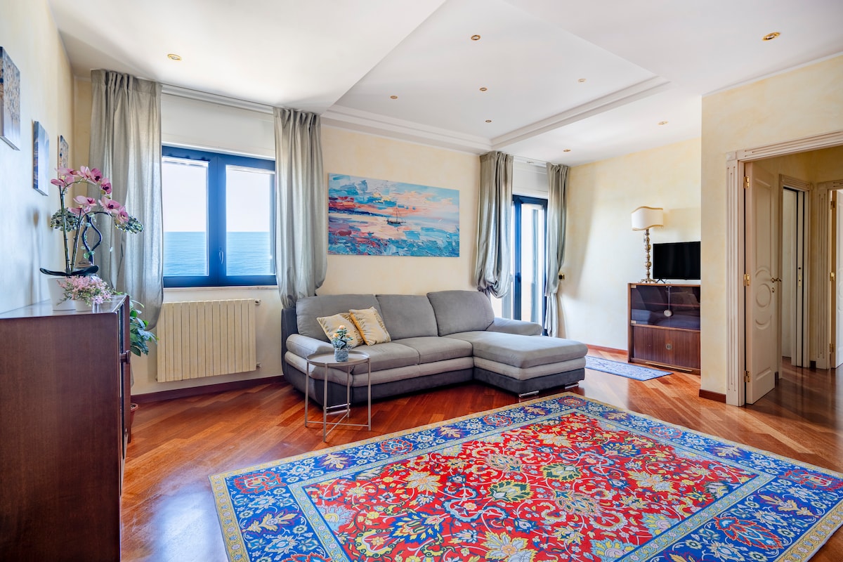 A spacious living room is highlighted by a soft grey sectional sofa and an ornate rug in vibrant colors. Large windows provide a view of the sea, while warm lighting enhances the tranquil environment. A small side table and a wooden cabinet are also visible.