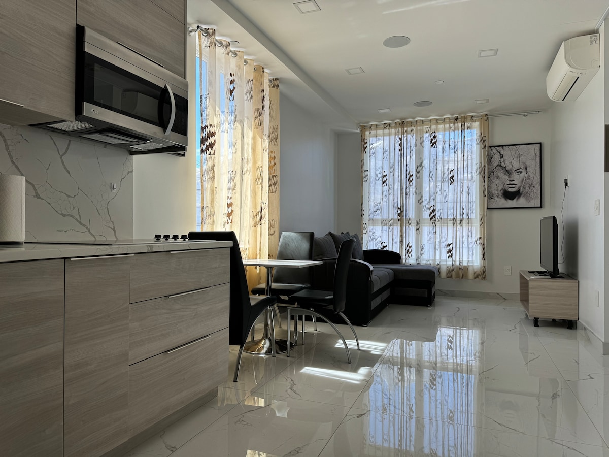 A modern kitchen area is paired with an open living space, showcasing sleek cabinetry and a stainless steel microwave. Natural light filters through sheer curtains, illuminating a comfortable couch and a small dining table. A flat-screen TV is mounted on a light-colored wall.