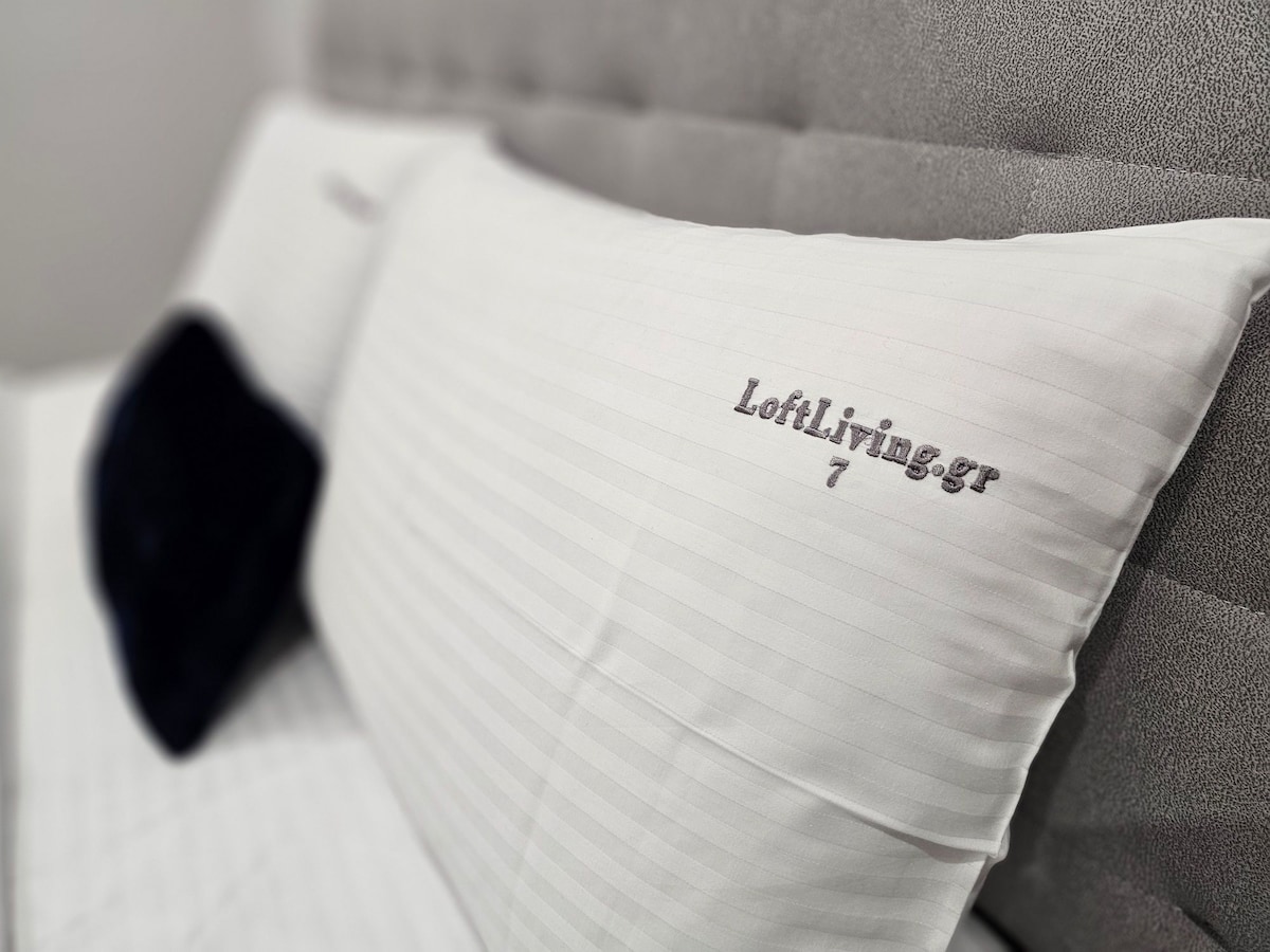 Two neatly arranged pillows on a plush, upholstered headboard are displayed. The pillows feature subtle striped patterns and the text 'LoftiLiving' is embroidered on them. A dark, velvety accent pillow adds a touch of contrast.