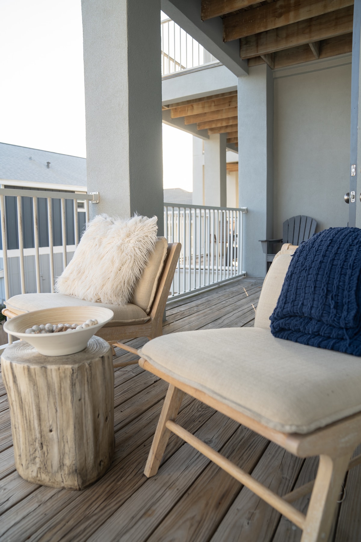 An outdoor seating area is presented with two comfortable chairs featuring neutral cushions. A rustic wooden stool holds a decorative bowl filled with small stones. Soft textiles are displayed on one chair, inviting a peaceful moment while overlooking the surroundings.