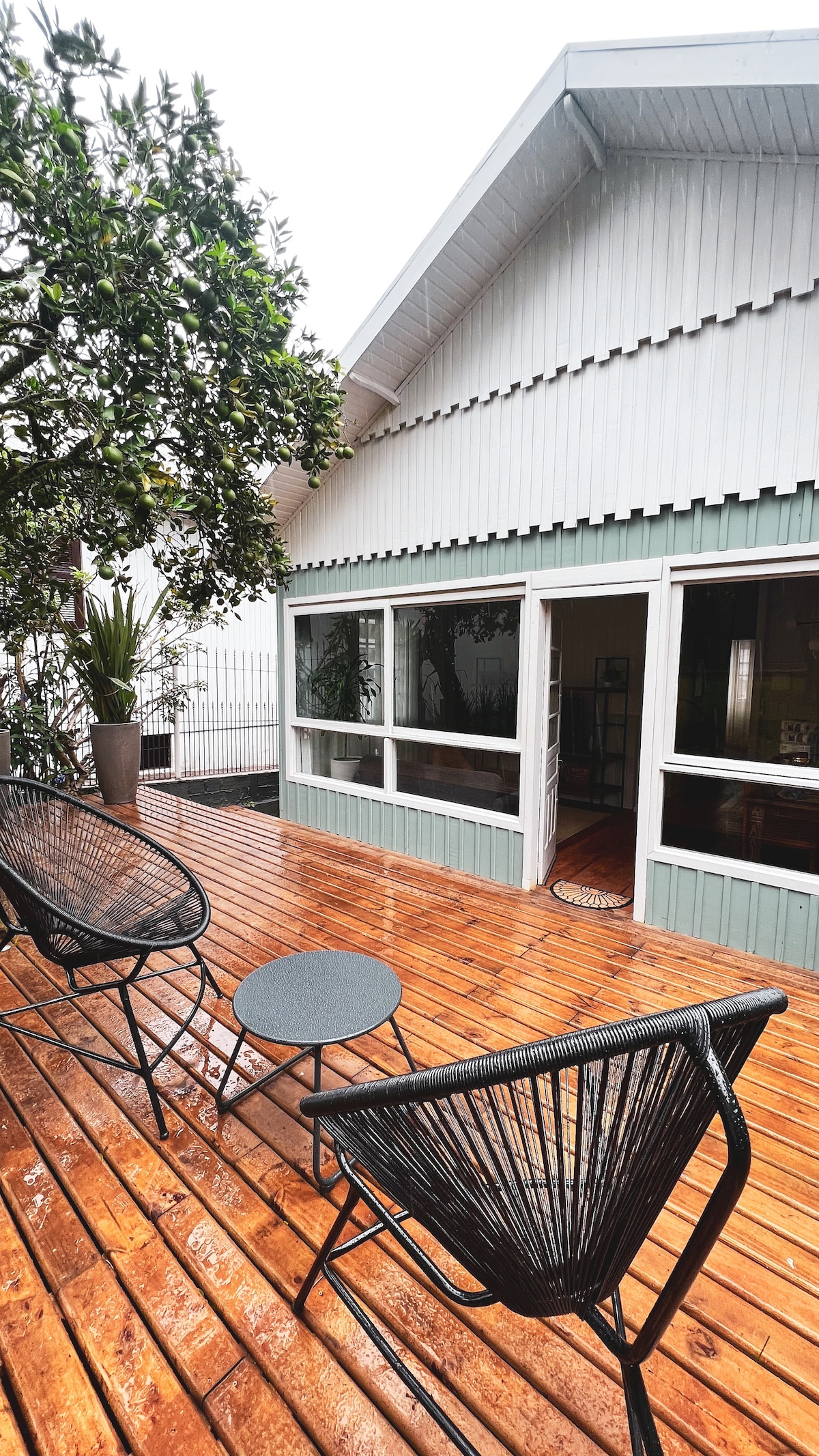 An open deck area features two black lounge chairs surrounding a small circular table. Large windows allow natural light to fill the interior space. The wooden flooring shows signs of recent rain, complementing the greenery from an adjacent tree.