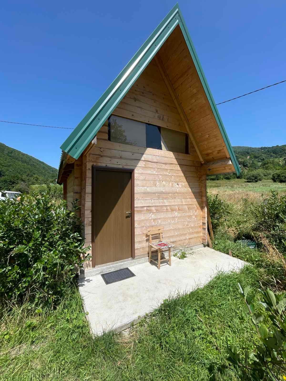 A wooden bungalow features a steep, green roof and large windows, allowing natural light to enter. A simple chair is placed on the concrete porch, surrounded by lush greenery and open fields, providing a connection to the serene natural surroundings.
