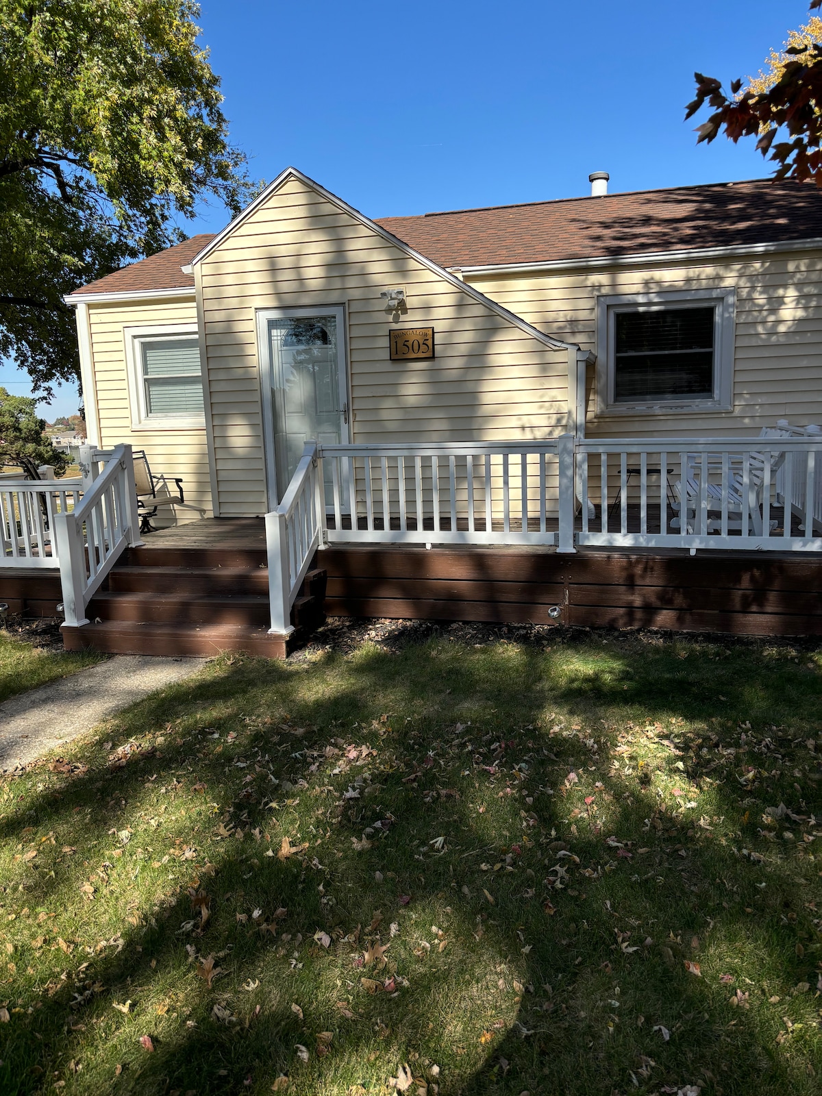 The exterior of a single-story house is shown, featuring a light-colored facade and a gabled roof. A welcoming front porch with white railings provides access to the front door. Surrounding greenery includes a well-maintained lawn under dappled sunlight.