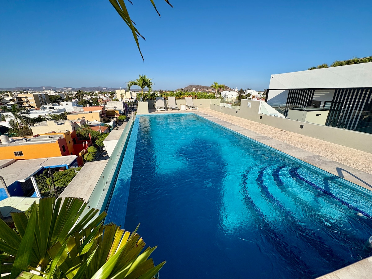 A rooftop pool is featured, showcasing clear blue water and surrounded by a stone deck. Lounge chairs are positioned on one side, with palm trees adding greenery in the foreground. The landscape offers a view of the nearby buildings and mountains under a clear blue sky.