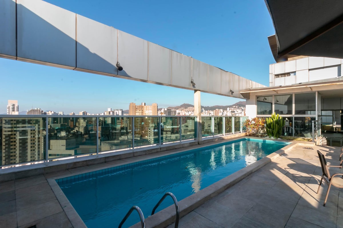A rooftop area features a swimming pool surrounded by a spacious terrace. Glass railings provide an unobstructed view of the city skyline. Lounge chairs are positioned nearby, enhancing the open and bright ambiance under clear blue skies.
