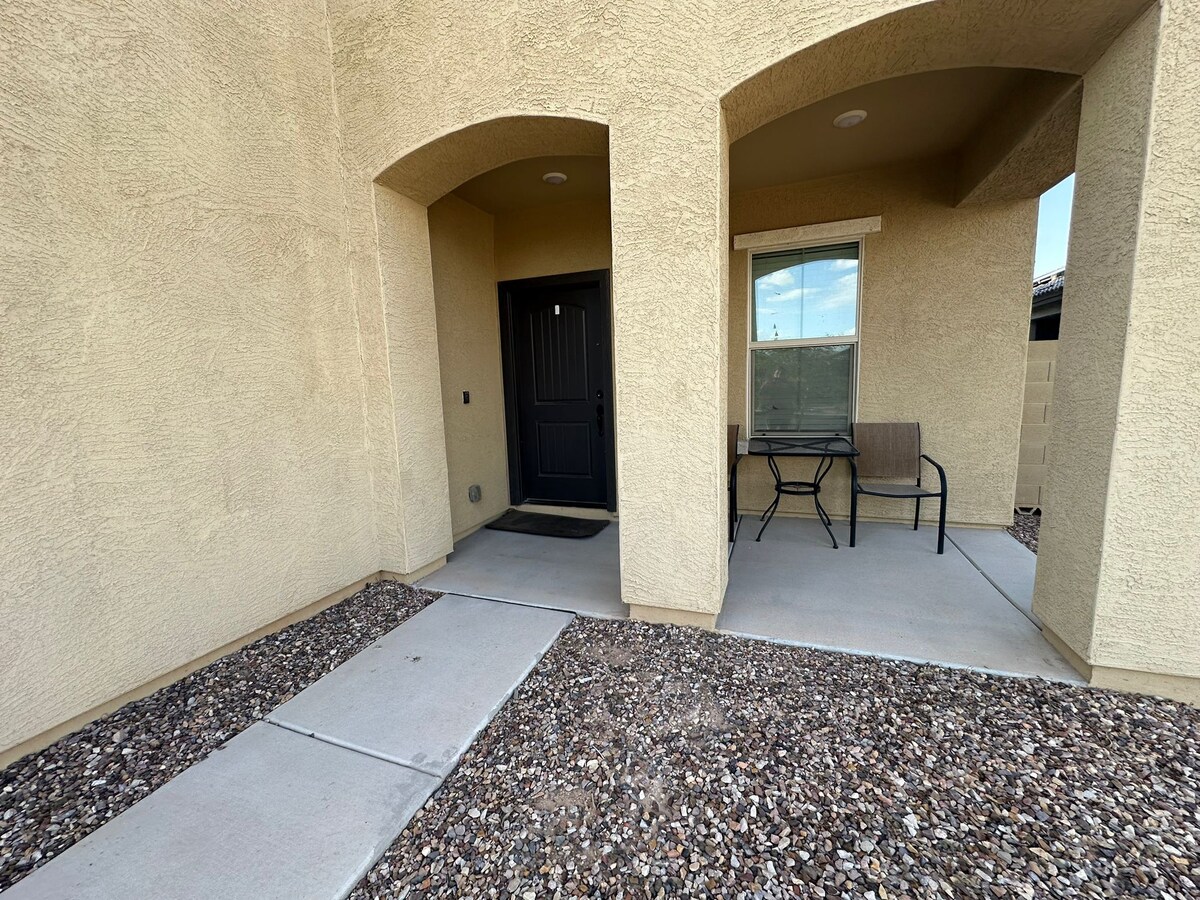 The entrance to the house features a covered porch with two simple chairs positioned on a concrete slab. The front door is black, set within a textured beige wall, with a window to the side allowing natural light.