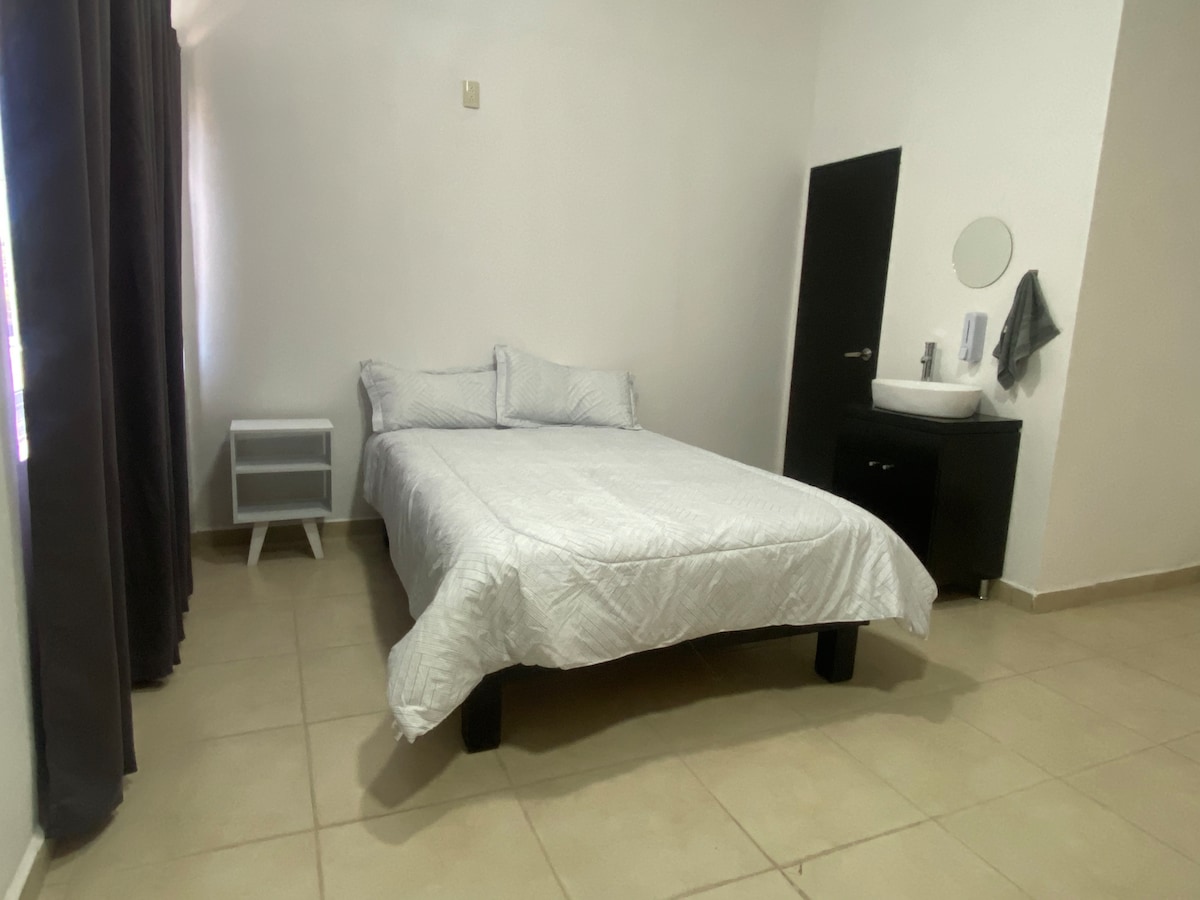 A bright sleeping area is furnished with a spacious bed covered in a quilted white duvet. A black nightstand stands beside the bed, and a circular mirror is mounted above a small sink. Dark curtains frame the window, allowing for a soft glow of natural light.