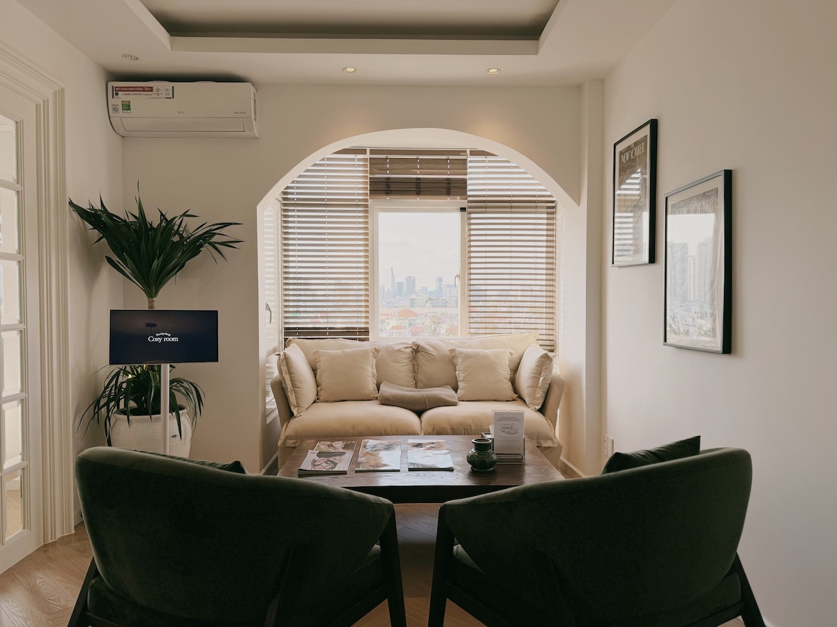 A cozy living area features a comfortable couch and two armchairs facing a coffee table. Natural light filters through large windows adorned with wooden blinds, offering views of the city skyline. Potted plants add a touch of greenery to the peaceful ambiance.