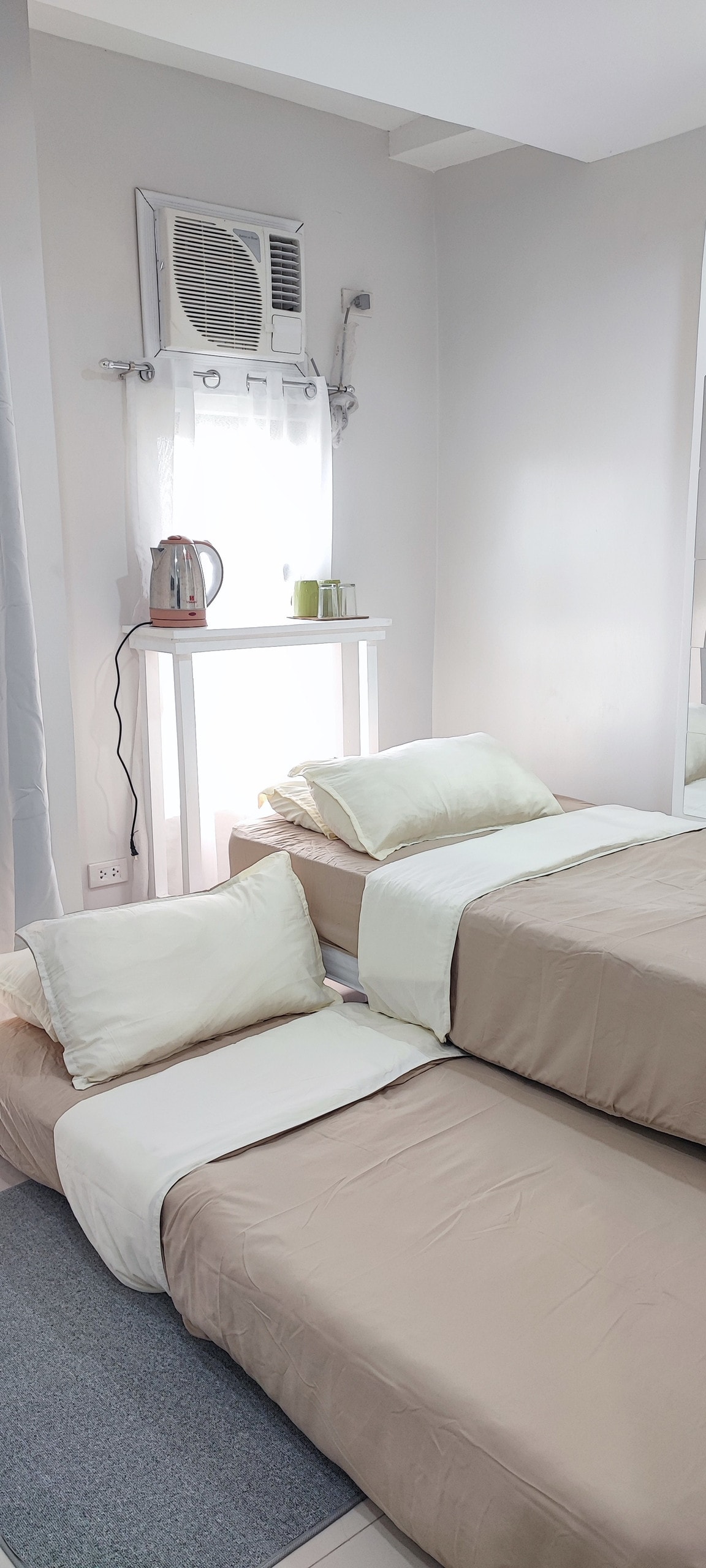 A comfortable sleeping area features two beds with light-colored bedding and soft pillows. A small electric kettle and cups are positioned on the shelf, while natural light enters through a nearby window with sheer curtains. The room is decorated in neutral tones.